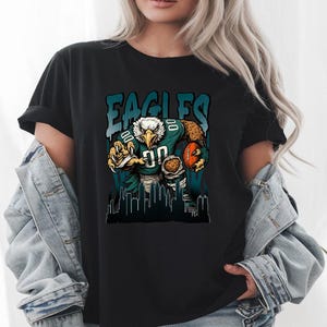 May include: Black t-shirt with a teal and white graphic of an eagle in a football uniform, holding a ball. The word "EAGLES" is printed above the eagle. A cityscape silhouette is at the bottom of the design.