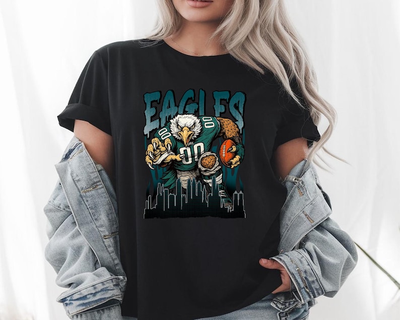 May include: Black t-shirt with a teal and white graphic of an eagle in a football uniform, holding a ball. The word "EAGLES" is printed above the eagle. A cityscape silhouette is at the bottom of the design.