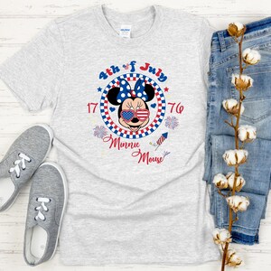 May include: A light gray t-shirt featuring a Minnie Mouse graphic with patriotic colors and the text "4th of July." The shirt is paired with blue jeans and gray sneakers, creating a casual, festive ensemble.