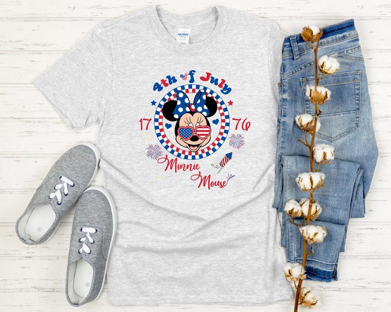 May include: A light gray t-shirt featuring a Minnie Mouse graphic with patriotic colors and the text "4th of July." The shirt is paired with blue jeans and gray sneakers, creating a casual, festive ensemble.