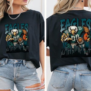May include: Black t-shirt with a graphic of an eagle in a football uniform holding a football. The word "EAGLES" is above the eagle in teal letters. A cityscape silhouette is at the bottom of the graphic.