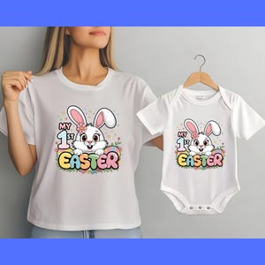 May include: White t-shirt with a graphic of a bunny holding an Easter egg and the text "My 1st Easter". A white baby onesie with the same graphic is also shown.