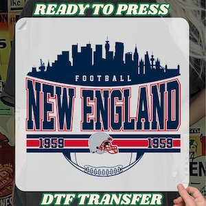 May include: A white DTF transfer featuring a navy blue skyline, the words "FOOTBALL NEW ENGLAND" in red and blue, and a football helmet. The transfer also includes the year "1959" and the text "READY TO PRESS" and "DTF TRANSFER".
