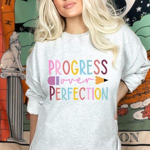 May include: A light gray sweatshirt with the words "PROGRESS over PERFECTION" in colorful letters and a pencil graphic. The sweatshirt is long-sleeved and has a crew neck.