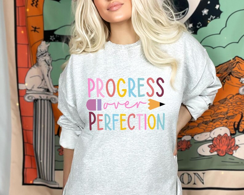 May include: A light gray sweatshirt with the words "PROGRESS over PERFECTION" in colorful letters and a pencil graphic. The sweatshirt is long-sleeved and has a crew neck.