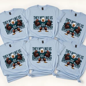 K&ouml;nnte beinhalten: Hellblaue T-Shirts mit einer Grafik eines muskul&ouml;sen Adlers, der ein Football-Trikot tr&auml;gt. Der Text auf den Shirts lautet "THEY NOT LIKE US BIRD GANG PHILADELPHIA FOOTBALL" und "01".