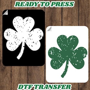 May include: Two rectangular DTF transfer designs featuring a distressed shamrock. One is a white shamrock on a black background, and the other is a green shamrock on a white background. The text "READY TO PRESS" and "DTF TRANSFER" are displayed in green.
