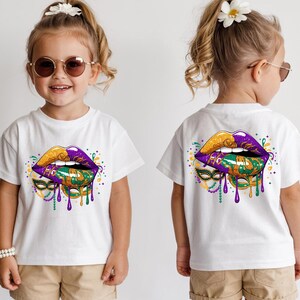 May include: White t-shirt featuring a vibrant graphic of stylized lips in gold, purple, and green, with dripping accents and a mask design. The design is on the front and back of the shirt. The shirt is worn by a child.