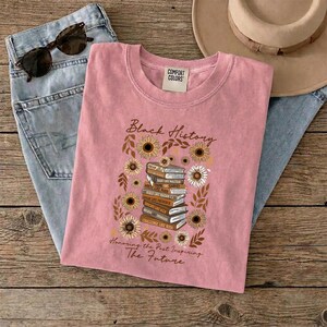 May include: A pink t-shirt with the words "Black History" and "Honoring the Past Inspiring The Future" printed on it. The design includes a stack of books, sunflowers, and leaves. The shirt is laid on top of blue jeans, sunglasses, and a hat.