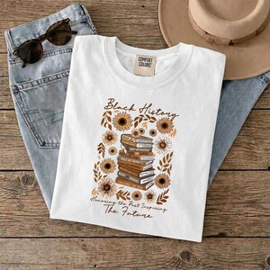 May include: White crew neck t-shirt with a graphic design featuring a stack of books, sunflowers, and the text "Black History." The design is in shades of brown and gold. The text "Honoring the Past Inspiring The Future" is also included.