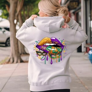 May include: A light gray hoodie featuring a vibrant graphic of lips in gold, purple, and green, with a matching mask and dripping accents. The design is centered on the back of the hoodie.