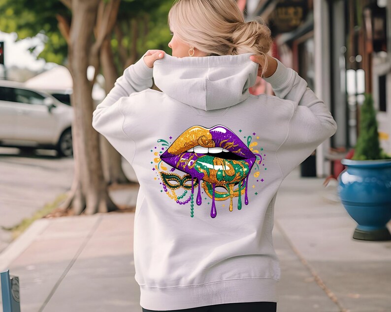 May include: A light gray hoodie featuring a vibrant graphic of lips in gold, purple, and green, with a matching mask and dripping accents. The design is centered on the back of the hoodie.