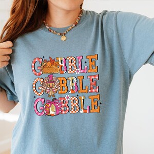 May include: A light blue t-shirt with the word "GOBBLE" in large, colorful letters. The letters are decorated with images of a pie, turkey, and pumpkin. The shirt has short sleeves and a crew neck.