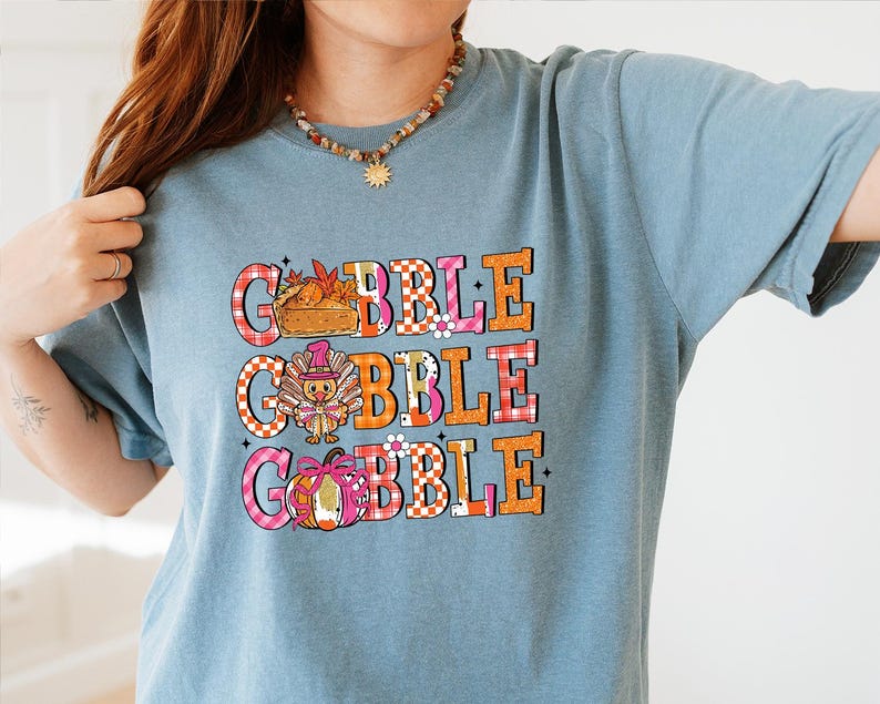 May include: A light blue t-shirt with the word "GOBBLE" in large, colorful letters. The letters are decorated with images of a pie, turkey, and pumpkin. The shirt has short sleeves and a crew neck.