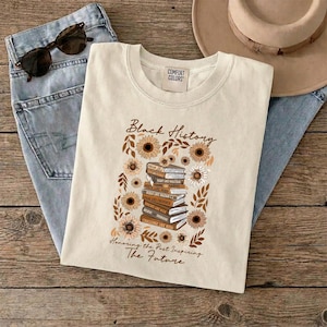May include: Cream-colored t-shirt with the words "Black History" and a floral design featuring sunflowers and books. The shirt also has the text "Honoring the Past Inspiring The Future". Sunglasses and jeans are also in the image.