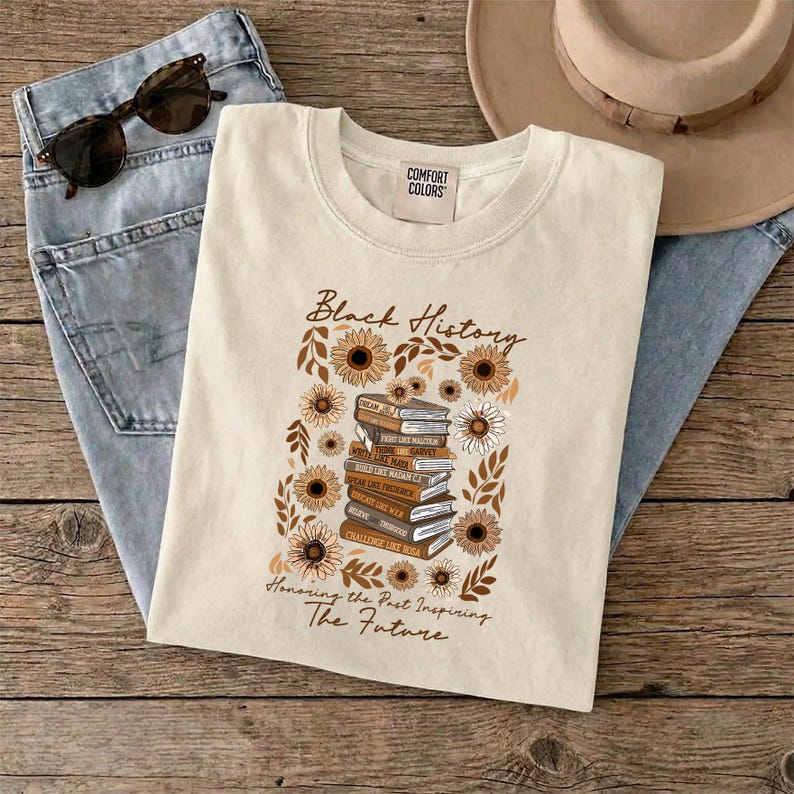May include: Cream-colored t-shirt with the words "Black History" and a floral design featuring sunflowers and books. The shirt also has the text "Honoring the Past Inspiring The Future". Sunglasses and jeans are also in the image.