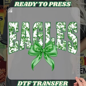May include: A green and white floral design with the word "EAGLES" in a bold font. A green bow is centred below the word. The design is on a clear background with the text "READY TO PRESS" and "DTF TRANSFER" at the top and bottom.