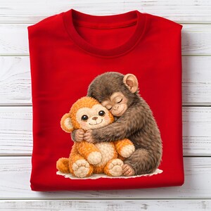 Op de afbeelding: Een rode crewneck sweatshirt met een afbeelding van een bruine aap die een oranje knuffel aap omhelst. De illustratie is in een zachte aquarel stijl. De apen worden afgebeeld met gesloten ogen, wat slaap of comfort suggereert.