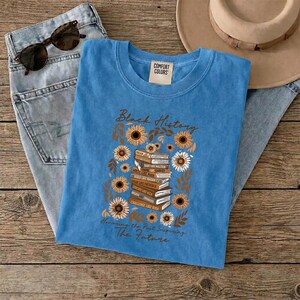 May include: A blue Comfort Colors t-shirt with the words "Black History" and "Honoring the Past, Inspiring The Future." The shirt features a stack of books surrounded by sunflowers and leaves. Sunglasses and jeans are also visible.