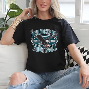 Może przedstawiać: Czarna koszulka z grafiką futbolu amerykańskiego Philadelphia Eagles. Projekt przedstawia orła trzymającego piłkę do futbolu, z napisami "PHILADELPHIA FOOTBALL EST 1933" w kolorze morskim i białym.
