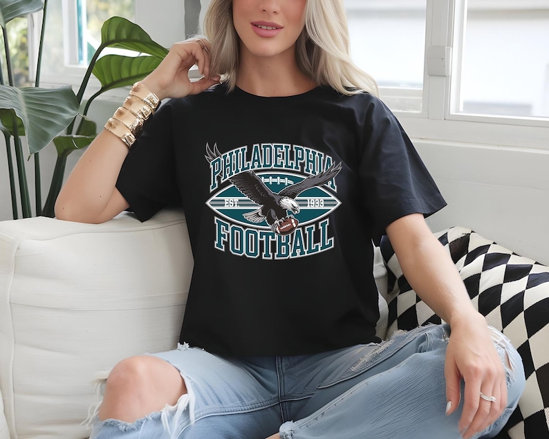 Może przedstawiać: Czarna koszulka z grafiką futbolu amerykańskiego Philadelphia Eagles. Projekt przedstawia orła trzymającego piłkę do futbolu, z napisami "PHILADELPHIA FOOTBALL EST 1933" w kolorze morskim i białym.
