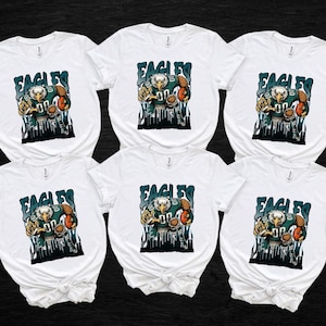 May include: Six white t-shirts featuring a graphic of an eagle in a football uniform holding a football. The word "EAGLES" is above the eagle in teal letters. The eagle graphic has teal, brown, and white colours.