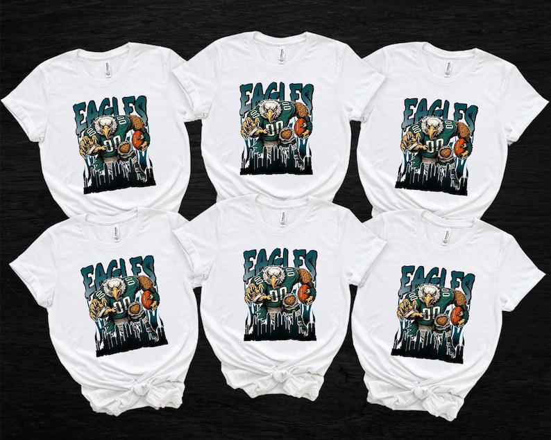 May include: Six white t-shirts featuring a graphic of an eagle in a football uniform holding a football. The word "EAGLES" is above the eagle in teal letters. The eagle graphic has teal, brown, and white colours.