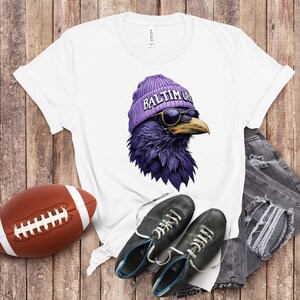 Pu&ograve; includere: T-shirt bianco con un corvo viola che indossa un berretto viola con la scritta "Baltimore" e occhiali da sole. Un pallone da football americano marrone, scarpe nere con lacci bianchi e jeans sono anche nell'immagine. La maglietta &egrave; su uno sfondo di legno.