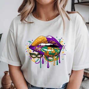 May include: White t-shirt featuring a vibrant graphic of lips in gold, purple, and green, with a matching mask design. The design includes dripping paint and beaded accents, evoking a festive Mardi Gras theme.