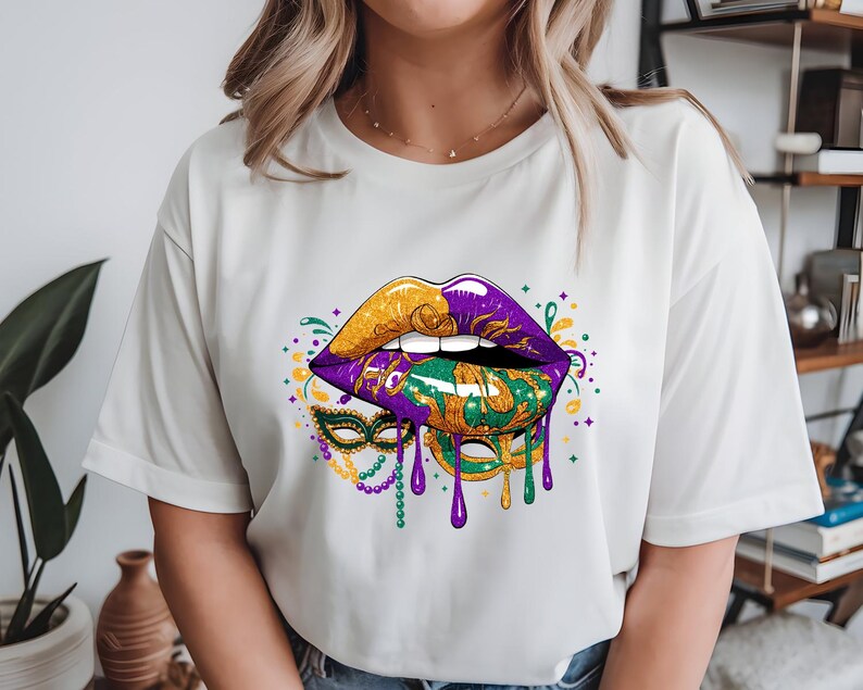 May include: White t-shirt featuring a vibrant graphic of lips in gold, purple, and green, with a matching mask design. The design includes dripping paint and beaded accents, evoking a festive Mardi Gras theme.