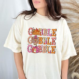May include: Cream-colored t-shirt with the text "GOBBLE GOBBLE" in a colorful, Thanksgiving-themed design. The design includes a turkey, pumpkin, and fall leaves. The shirt has short sleeves and a relaxed fit.