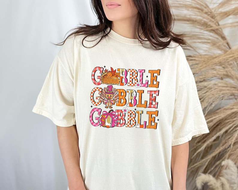 May include: Cream-colored t-shirt with the text "GOBBLE GOBBLE" in a colorful, Thanksgiving-themed design. The design includes a turkey, pumpkin, and fall leaves. The shirt has short sleeves and a relaxed fit.