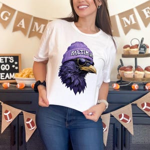 Pu&ograve; includere: T-shirt bianca con una grafica di un corvo viola che indossa un berretto viola con la scritta "BALTIMORE" e occhiali da sole. Il corvo &egrave; un'illustrazione dettagliata. La maglietta &egrave; in stile casual.