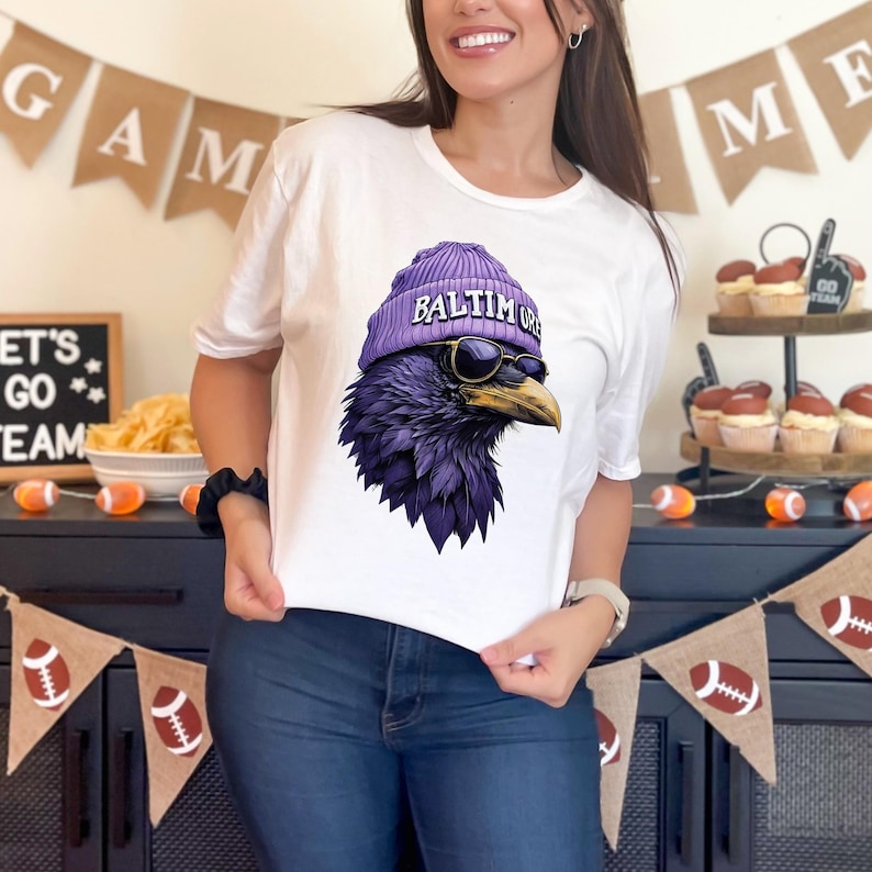 Pu&ograve; includere: T-shirt bianca con una grafica di un corvo viola che indossa un berretto viola con la scritta "BALTIMORE" e occhiali da sole. Il corvo &egrave; un'illustrazione dettagliata. La maglietta &egrave; in stile casual.