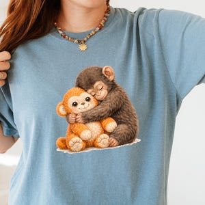 Op de afbeelding: Lichtblauw T-shirt met een afbeelding van twee knuffelende apen. E&eacute;n aap is bruin, de andere oranje. De apen knuffelen elkaar. Het T-shirt is gemaakt van een zacht materiaal.