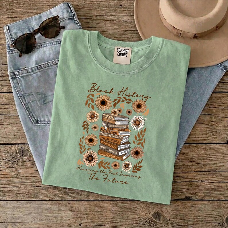 May include: A sage green t-shirt with the words "Black History" and "Honoring the Past Inspiring The Future". The graphic features a stack of books surrounded by sunflowers and leaves. Sunglasses and jeans are also in the image.