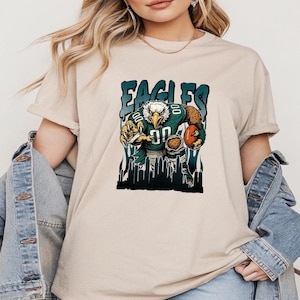 May include: Beige t-shirt with a graphic of an eagle in a football uniform, holding a football. The word "EAGLES" is printed above the eagle in a teal font. The eagle is set against a cityscape background.
