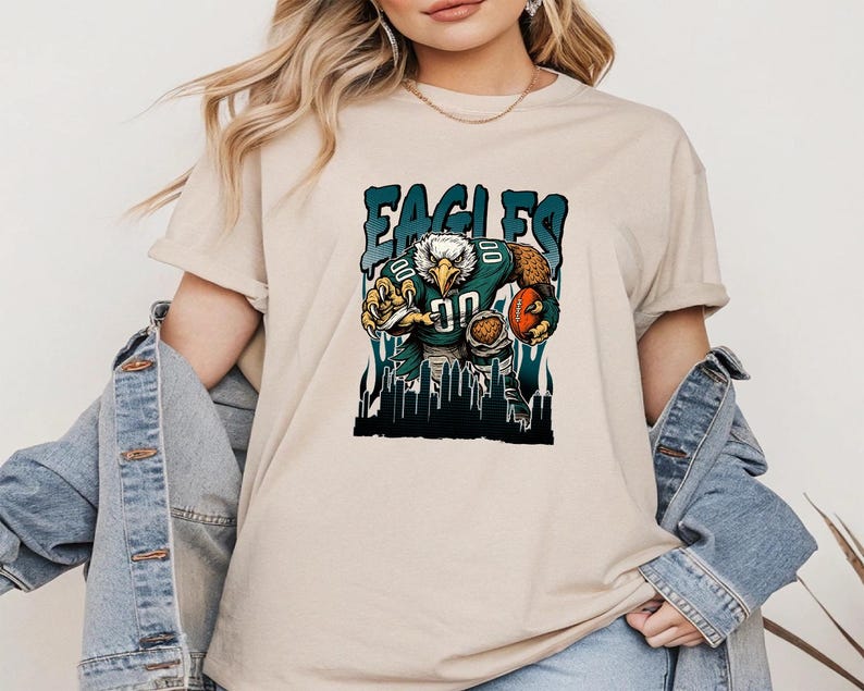 May include: Beige t-shirt with a graphic of an eagle in a football uniform, holding a football. The word "EAGLES" is printed above the eagle in a teal font. The eagle is set against a cityscape background.