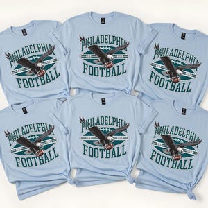 Może przedstawiać: Jasnoniebieskie koszulki z nadrukiem Philadelphia Football. Projekt przedstawia piłkę nożną, orła i napisy "Philadelphia Football" w kolorze białym i ciemnozielonym. Koszulki są z kr&oacute;tkim rękawem.