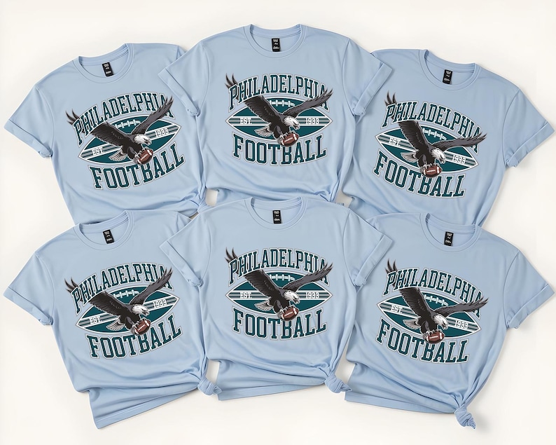 Może przedstawiać: Jasnoniebieskie koszulki z nadrukiem Philadelphia Football. Projekt przedstawia piłkę nożną, orła i napisy "Philadelphia Football" w kolorze białym i ciemnozielonym. Koszulki są z kr&oacute;tkim rękawem.