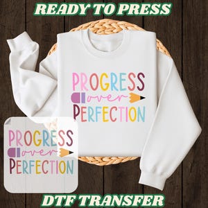 May include: White sweatshirt with the text "PROGRESS over PERFECTION" in colorful letters and a pencil graphic. The words "READY TO PRESS" and "DTF TRANSFER" are also displayed. The sweatshirt is on a woven mat against a wooden background.