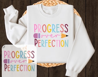 May include: White sweatshirt with the text "PROGRESS over PERFECTION" in colorful letters and a pencil graphic. The words "READY TO PRESS" and "DTF TRANSFER" are also displayed. The sweatshirt is on a woven mat against a wooden background.