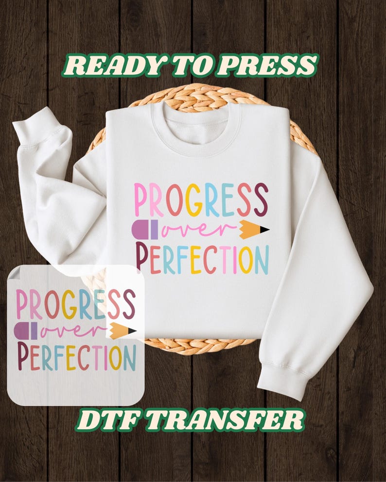 May include: White sweatshirt with the text "PROGRESS over PERFECTION" in colorful letters and a pencil graphic. The words "READY TO PRESS" and "DTF TRANSFER" are also displayed. The sweatshirt is on a woven mat against a wooden background.