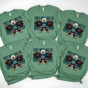 K&ouml;nnte beinhalten: Sechs gr&uuml;ne T-Shirts mit dem Text "THEY NOT LIKE US BIRD GANG PHILADELPHIA FOOTBALL" und einem Cartoon-Adler, der ein Football-Trikot tr&auml;gt. Der Adler spannt seine Muskeln an. Die Hemden sind gefaltet.