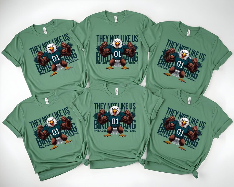 K&ouml;nnte beinhalten: Sechs gr&uuml;ne T-Shirts mit dem Text "THEY NOT LIKE US BIRD GANG PHILADELPHIA FOOTBALL" und einem Cartoon-Adler, der ein Football-Trikot tr&auml;gt. Der Adler spannt seine Muskeln an. Die Hemden sind gefaltet.