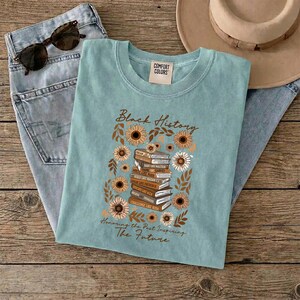 May include: A light blue t-shirt with the words "Black History" and "Honoring the Past Inspiring The Future". The shirt features a graphic of sunflowers and a stack of books. Sunglasses and jeans are in the background.
