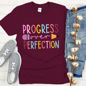 May include: A maroon t-shirt with the words "PROGRESS over PERFECTION" in colorful letters and a pencil graphic. The shirt is paired with blue jeans and gray sneakers.