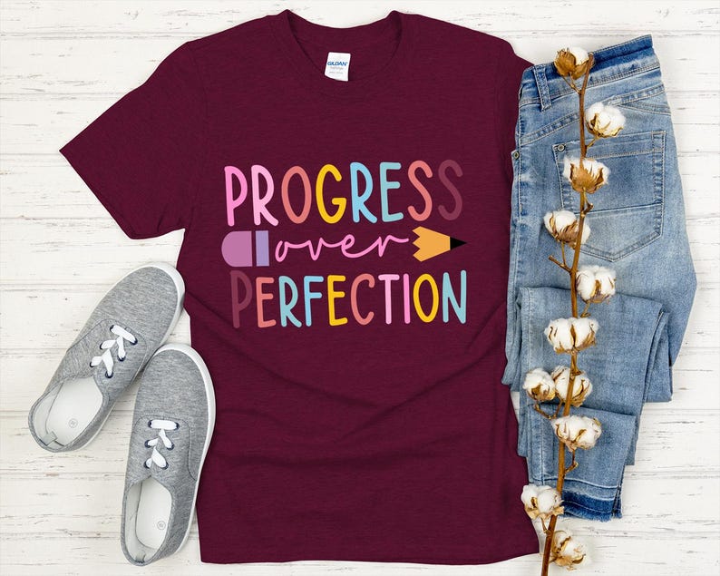 May include: A maroon t-shirt with the words "PROGRESS over PERFECTION" in colorful letters and a pencil graphic. The shirt is paired with blue jeans and gray sneakers.