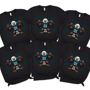 K&ouml;nnte beinhalten: Sechs schwarze T-Shirts mit einem Cartoon-Adler-Design. Der Adler spannt seine Muskeln an und tr&auml;gt ein t&uuml;rkisfarbenes Trikot mit der Nummer 01. Der Adler ist vor einem t&uuml;rkisfarbenen Hintergrund platziert. Die Shirts sind kurz&auml;rmelig.