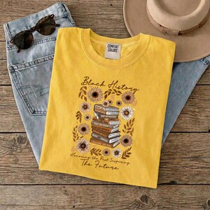 May include: Yellow t-shirt with the words "Black History" and a graphic of books and sunflowers. The shirt also has the text "Honoring the Past Inspiring The Future". The shirt is folded on top of blue jeans, sunglasses, and a hat.